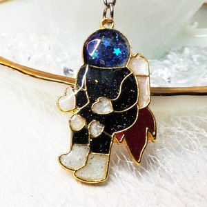 Puede incluir: Un llavero dorado con una figura de astronauta de resina con un casco espacial azul y negro. El astronauta está sentado en un cohete rojo con detalles blancos.