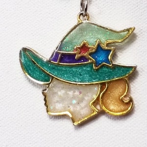 May include: A green and gold witch charm with glitter accents. The charm features a witch with a green hat and a white face. The charm is attached to a silver chain with a green bead.