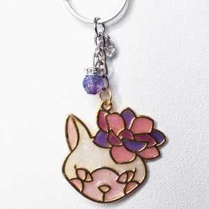 Puede incluir: Llavero con diseño de cara de gato en blanco y rosa, adornado con una corona de flores rosas y moradas y bordes dorados. Una pequeña cuenta de purpurina morada y una cadena plateada completan el diseño.