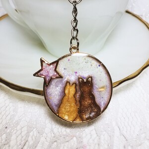 Puede incluir: Un llavero dorado con dos gatos sentados espalda con espalda bajo una estrella rosa. Los gatos están delineados en negro y tienen un fondo blanco brillante.