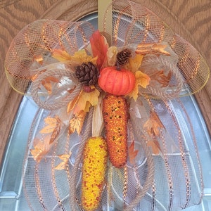 May include: A fall-themed door wreath with a brown burlap bow, orange and yellow corn, a small orange pumpkin, and gold leaves.