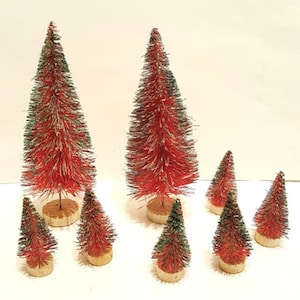 May include: A collection of miniature bottle brush Christmas trees. The trees are red with green tips and are mounted on small wooden bases. The trees vary in size and are arranged on a white surface.