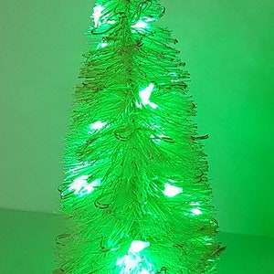 May include: A small, white, artificial Christmas tree with green lights. The tree is decorated with a string of lights that are lit up.