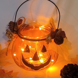 May include: A glowing orange glass jack-o'-lantern with a black handle and a smiling face. The Halloween decoration is adorned with pinecones, berries, and a string of lights. Autumn leaves are scattered around the base.