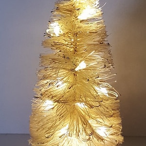 May include: A small, white, brush-style artificial Christmas tree with warm white LED lights. The tree is lit and sitting on a wooden base.