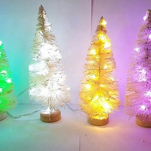 May include: Six white artificial Christmas trees with lights. The trees are decorated with different coloured lights, including blue, green, white, yellow, purple, and orange.