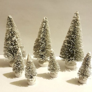 May include: A set of seven white flocked artificial Christmas trees of varying sizes. The trees are arranged on a white surface.