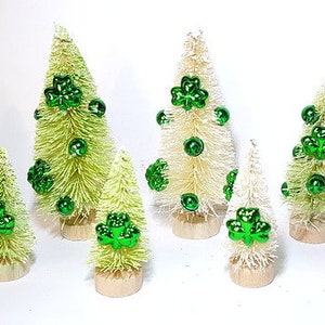 May include: Seven miniature white bottle brush trees decorated with green shamrock ornaments. The trees are of varying sizes and are arranged on a white surface.