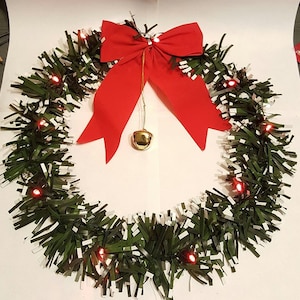 Puede incluir: Una corona de Navidad verde y blanca con luces rojas y un lazo rojo con una campana dorada colgando.