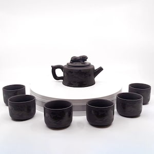 Op de afbeelding: Een donkerbruine theepot met een klein dierlijk figuurtje op de deksel, omringd door zes bijpassende theekopjes op een wit oppervlak.