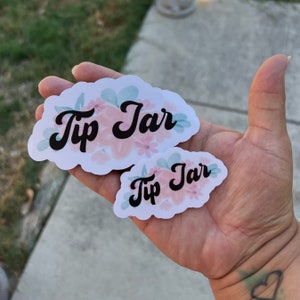 May include: Two white stickers with pink and blue floral designs. The larger sticker says "Tip Jar" in black cursive font. The smaller sticker says "Tip Jar" in black cursive font.