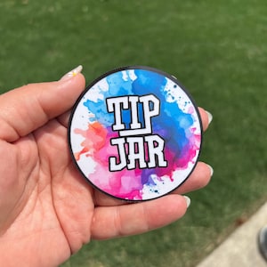 May include: A round sticker with a white background and colorful watercolor splatters in shades of pink, blue, and purple. The text "TIP JAR" is printed in black letters.