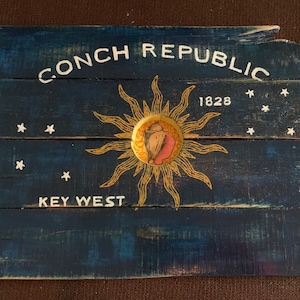 May include: A rustic, wooden sign painted dark blue, featuring the words "CONCH REPUBLIC" and "KEY WEST" in white. A sunburst design surrounds a shell emblem. The year "1828" is also displayed. White stars are scattered across the sign.