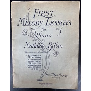 Puede incluir: Portada de partituras antiguas, "First Melody Lessons for Piano" de Mathilde Bilbro. El diseño incluye motivos florales y letras azul oscuro. Publicado por Hatch Music Company, Boston. El papel tiene un aspecto vintage.