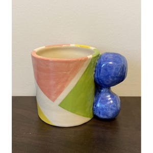 May include: A ceramic mug with a blue figure handle. The mug's exterior features a geometric design with pink, green, and yellow sections. The interior is a cream color. The mug is on a dark brown surface.