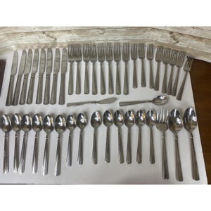 May include: A collection of stainless steel silverware, including knives, forks, spoons, and butter knives. The silverware is arranged on a white surface, with a wooden background.
