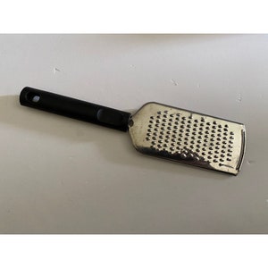Puede incluir: Un rallador de plata con un mango de plástico negro. El rallador tiene una malla fina y está diseñado para rallar queso u otros alimentos.