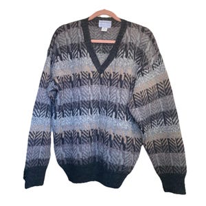 Könnte beinhalten: Ein V-Ausschnitt-Pullover in Grau-, Schwarz- und Beigetönen. Der Pullover hat ein strukturiertes, gemustertes Design mit geripptem Saum und Bündchen. Das Kleidungsstück hängt an einem rosa Kleiderbügel vor weißem Hintergrund.