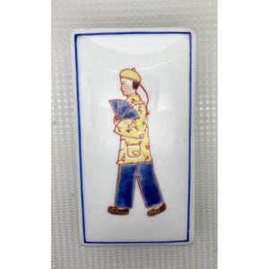 以下が含まれることがあります： 青い縁取りのある長方形の白いセラミック製の小物入れ。蓋には、扇子を持った伝統的な衣装を着た人物のカラフルなイラストが描かれています。その人物は、黄色のコート、青いズボン、黄色の帽子を身に着けています。