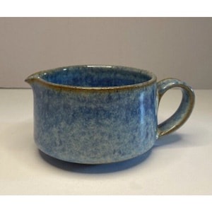 May include: A blue ceramic gravy boat with a brown rim and handle. The boat has a speckled glaze.