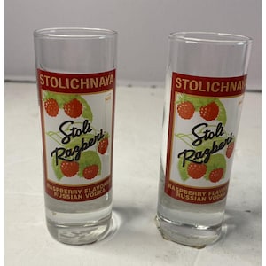以下が含まれることがあります： 赤いラベルと白いラベルがついた透明なガラス製のショットグラスが2つ。ラベルには「Stolichnaya Stoli Razberi Raspberry Flavored Russian Vodka」と書いてあります。