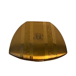 May include: A gold-toned, art deco style compact with a unique shape. The compact features vertical striped detailing and ornate floral accents. The initials "MW" are elegantly scripted in the center. The compact is closed and appears to be in good condition.