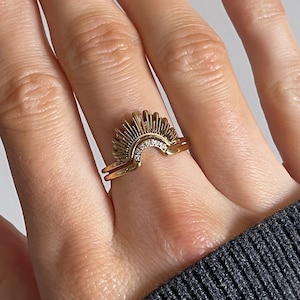 May include: A gold ring set with a sunburst design and small, clear stones. The ring is worn on a hand with manicured nails. The design includes a smaller band. The background is neutral. The ring's design is reminiscent of a sunrise.