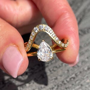 May include: A gold ring set with a pear-cut diamond and a wave-shaped band set with small diamonds. The ring is held up, showing its intricate design. The background is a grey wooden surface.
