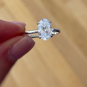 K&ouml;nnte beinhalten: Ein silberfarbener Ring mit einem einzelnen, klaren, oval geschliffenen Edelstein in einer Vier-Krappen-Fassung. Die Ringschiene ist schmal und poliert und weist eine reflektierende metallische Oberfl&auml;che auf, die den facettierten Stein betont.