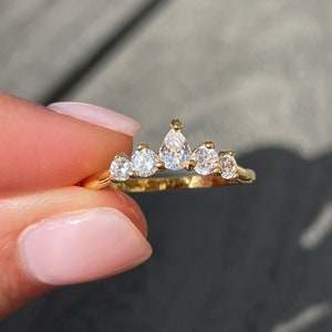 Peut inclure: Une bague en or avec cinq pierres transparentes à facettes disposées en arc de cercle. La pierre centrale est en forme de poire, flanquée de deux pierres rondes de chaque côté, le tout monté sur un anneau en métal doré incurvé au centre.
