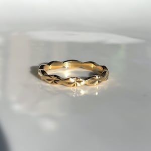 May include: A gold ring with a repeating starburst pattern. The ring is made of a shiny, yellow metal and is set against a white background. The ring's design features a series of diamond-shaped facets.