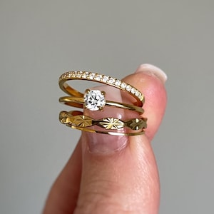 May include: Four gold rings are stacked together. The top ring features a curved design set with small, clear stones. Beneath it is a ring with a single, round, clear stone. The bottom ring has a repeating starburst pattern. The rings are held by a person's fingers.