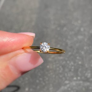 May include: A thin gold band ring featuring a single round brilliant-cut clear gemstone held in a four-prong setting. The metal band is smooth and polished, showcasing a minimalist design that highlights the central stone's clarity and sparkle.