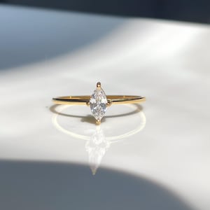 May include: A gold ring featuring a marquise-cut diamond. The ring has a thin band and a central diamond, creating a minimalist design. The ring is set on a white surface with shadows. The diamond's shape is elongated.
