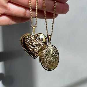 May include: Two gold-toned metal lockets on thin matching chains. One locket is heart-shaped with an embossed rose design, while the other is oval-shaped with an intricate floral pattern engraved across the entire surface of the metal.