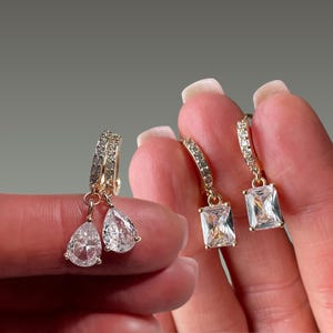Peut inclure: Une paire de boucles d'oreilles créoles dorées avec de petites pierres claires sur l'anneau et une pierre claire en forme de goutte, et une paire de créoles dorées avec des pierres claires et une pierre rectangulaire pendante.