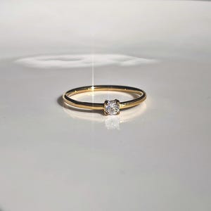 Peut inclure: Une fine bague en or avec une seule petite pierre précieuse ronde et transparente, montée sur un sertissage à quatre griffes. L'anneau en métal est lisse et poli, reflétant la lumière, tandis que la pierre centrale brille intensément.