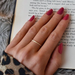 May include: A thin, gold-colored metal ring with a smooth, circular band. The ring is worn on the ring finger, which is positioned over an open book with printed text. The ring features a simple, unadorned design with a polished, reflective finish.