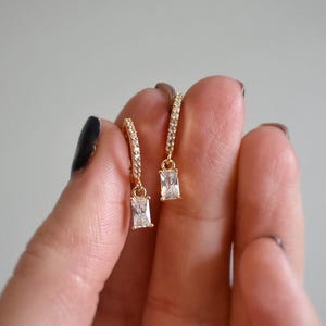 May include: Gold hoop earrings with a dangling rectangular clear stone. The earrings are set with small clear stones.