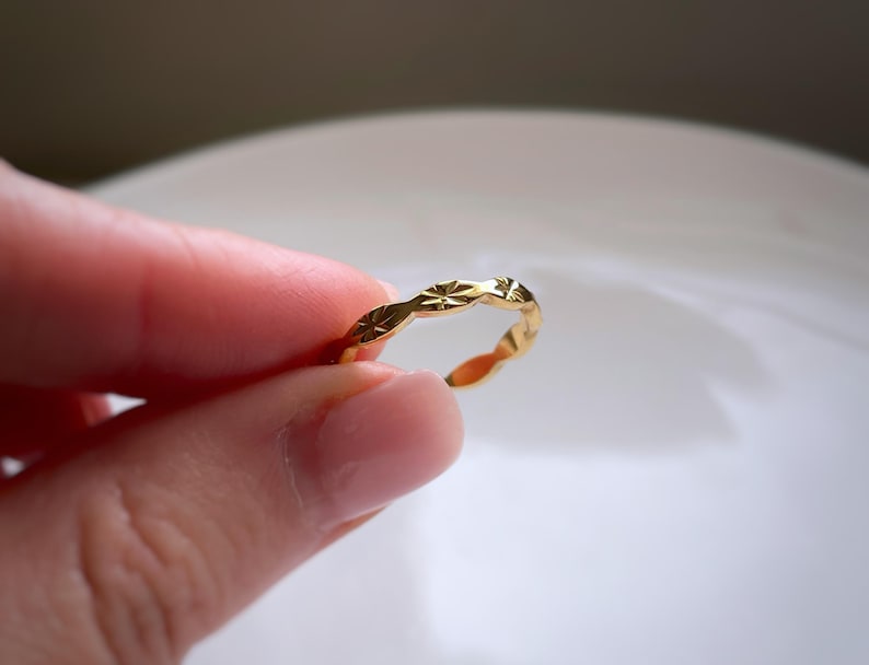 Peut inclure: Une bague dor&eacute;e pr&eacute;sentant une s&eacute;rie de motifs en &eacute;toile grav&eacute;s et r&eacute;p&eacute;t&eacute;s sur la bande ext&eacute;rieure. La surface m&eacute;tallique est polie et r&eacute;fl&eacute;chissante, avec un design &agrave; bords festonn&eacute;s qui suit le contour de chaque motif &eacute;toil&eacute;.