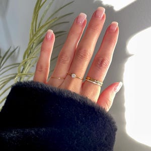 Peut inclure: Un ensemble de quatre bagues dor&eacute;es port&eacute;es aux doigts. Une bague pr&eacute;sente une pierre ronde transparente, une autre est un anneau fin et lisse, une troisi&egrave;me a un motif textur&eacute; et la derni&egrave;re est un anneau fin serti de petites pierres.
