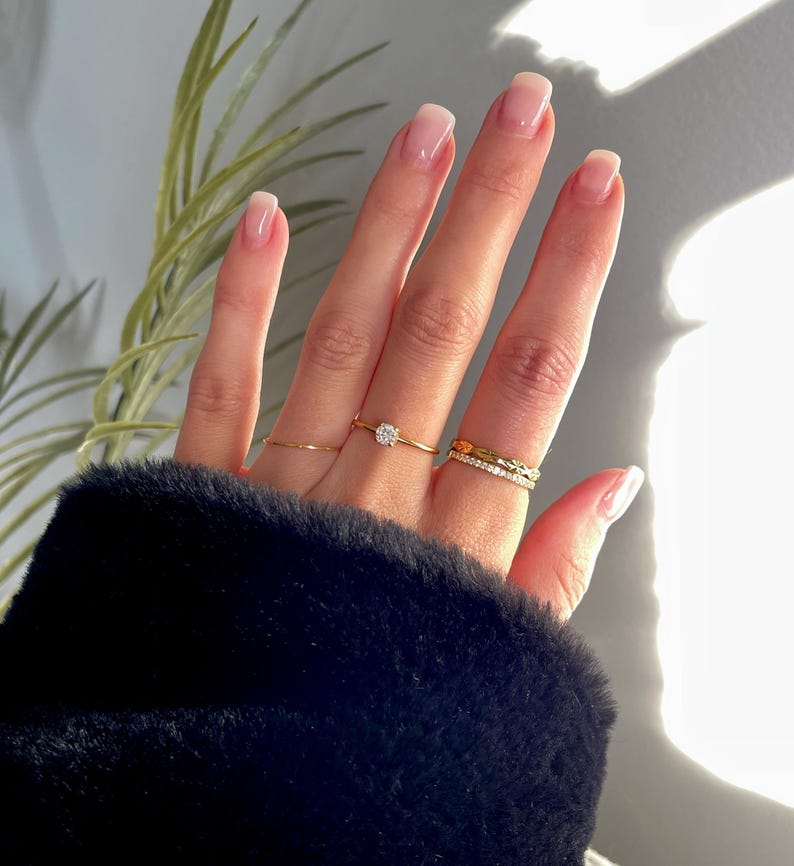 Peut inclure: Un ensemble de quatre bagues dor&eacute;es port&eacute;es aux doigts. Une bague pr&eacute;sente une pierre ronde transparente, une autre est un anneau fin et lisse, une troisi&egrave;me a un motif textur&eacute; et la derni&egrave;re est un anneau fin serti de petites pierres.