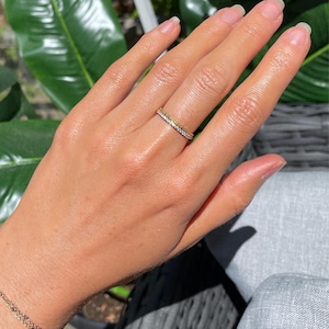May include: A thin gold band ring featuring a row of small, clear, sparkling stones set along the outer surface. The ring is worn on the ring finger, showcasing a metallic yellow finish and a delicate, uniform design with a polished texture.