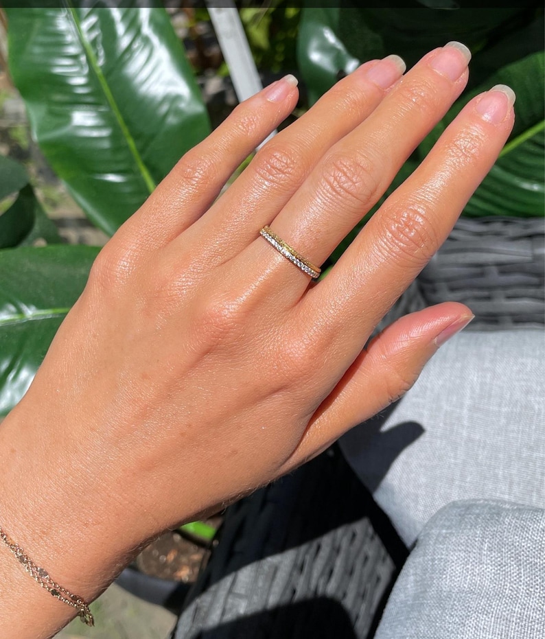 May include: A thin gold band ring featuring a row of small, clear, sparkling stones set along the outer surface. The ring is worn on the ring finger, showcasing a metallic yellow finish and a delicate, uniform design with a polished texture.