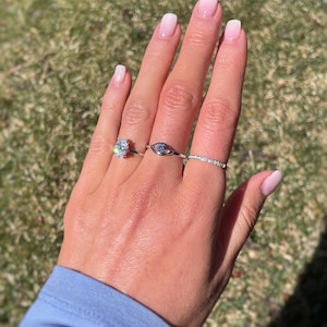 K&ouml;nnte beinhalten: Drei silberfarbene Ringe an den Fingern. Der erste Ring hat einen gro&szlig;en, oval geschliffenen, klaren Stein. Der zweite Ring ist ein augenf&ouml;rmiges Band mit einem kleinen blauen Stein in der Mitte. Der dritte Ring ist ein d&uuml;nnes, besetztes Band.