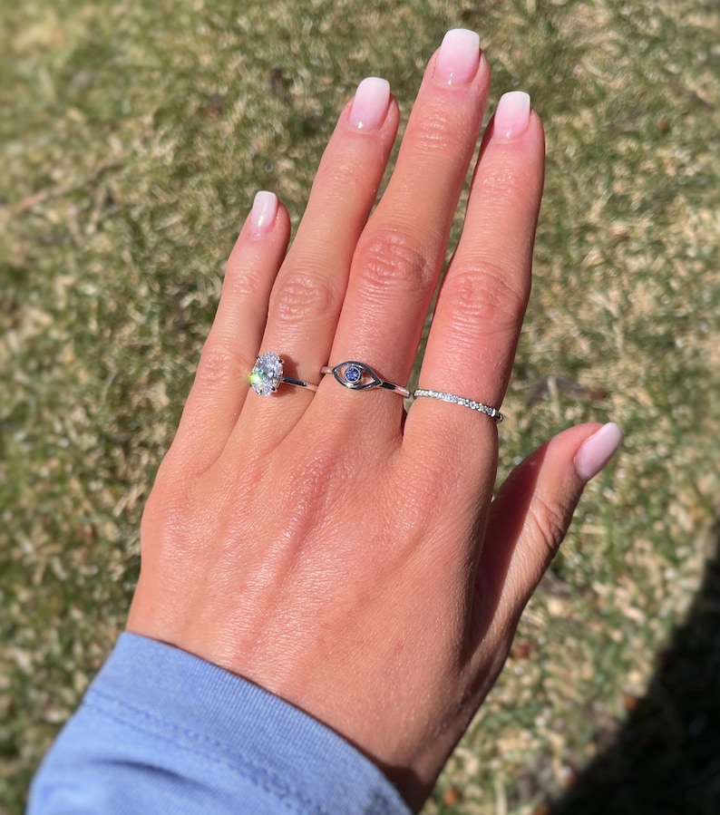 K&ouml;nnte beinhalten: Drei silberfarbene Ringe an den Fingern. Der erste Ring hat einen gro&szlig;en, oval geschliffenen, klaren Stein. Der zweite Ring ist ein augenf&ouml;rmiges Band mit einem kleinen blauen Stein in der Mitte. Der dritte Ring ist ein d&uuml;nnes, besetztes Band.