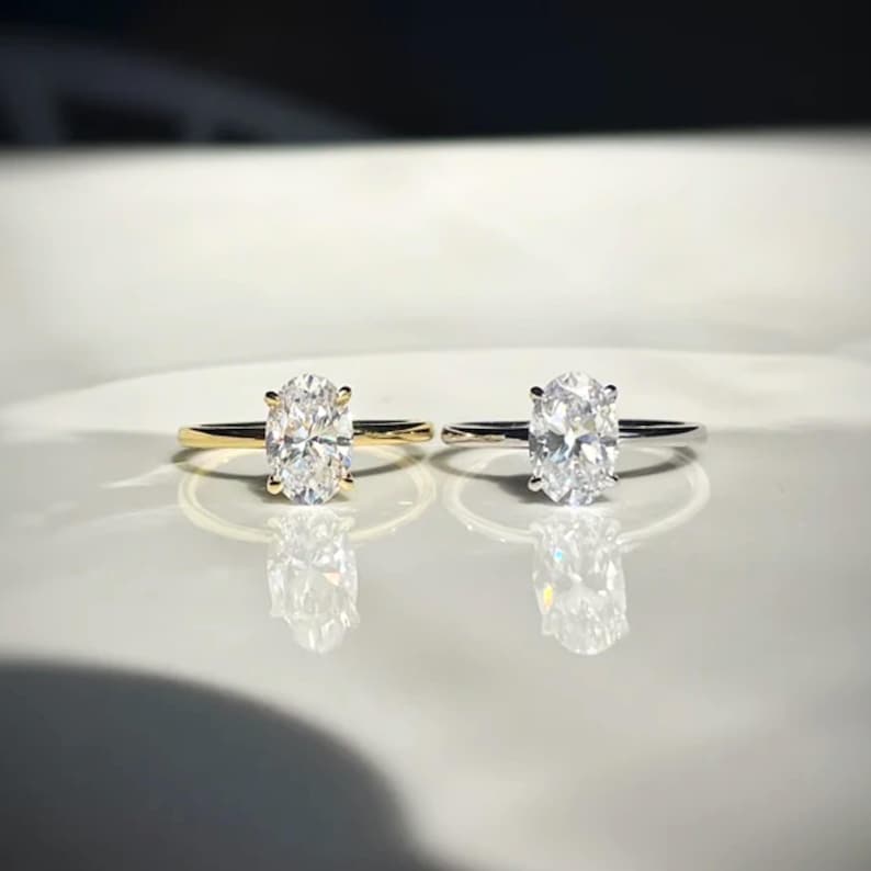 K&ouml;nnte beinhalten: Zwei Ringe mit oval geschliffenen, klaren Steinen in Vier-Krappen-Fassungen. Ein Ring hat eine Ringschiene aus Gelbgold, der andere eine silberfarbene Ringschiene. Beide Ringe stehen nebeneinander und reflektieren Licht auf einer glatten Fl&auml;che.