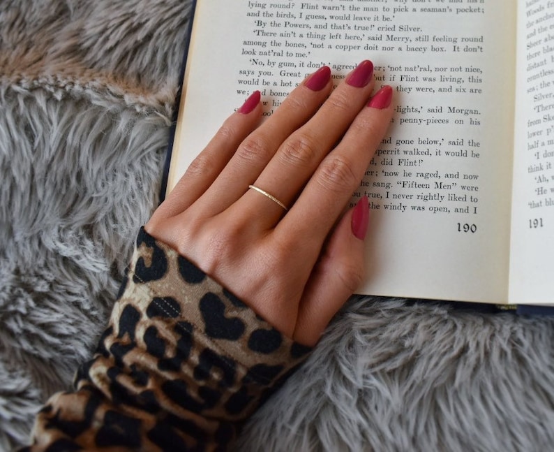 May include: A thin, gold-colored metal ring with a smooth, circular band. The ring is worn on the ring finger, which is positioned over an open book with printed text. The ring features a simple, unadorned design with a polished, reflective finish.