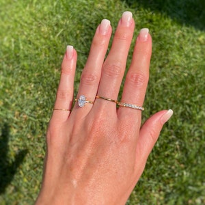 K&ouml;nnte beinhalten: Ein Set aus vier Goldringen an den Fingern. Ein Ring hat einen markanten oval geschliffenen klaren Stein, ein weiterer ist ein d&uuml;nner schlichter Goldring, der dritte ist ein einfacher Goldring und der vierte ist ein mit kleinen klaren Steinen besetzter Ring.
