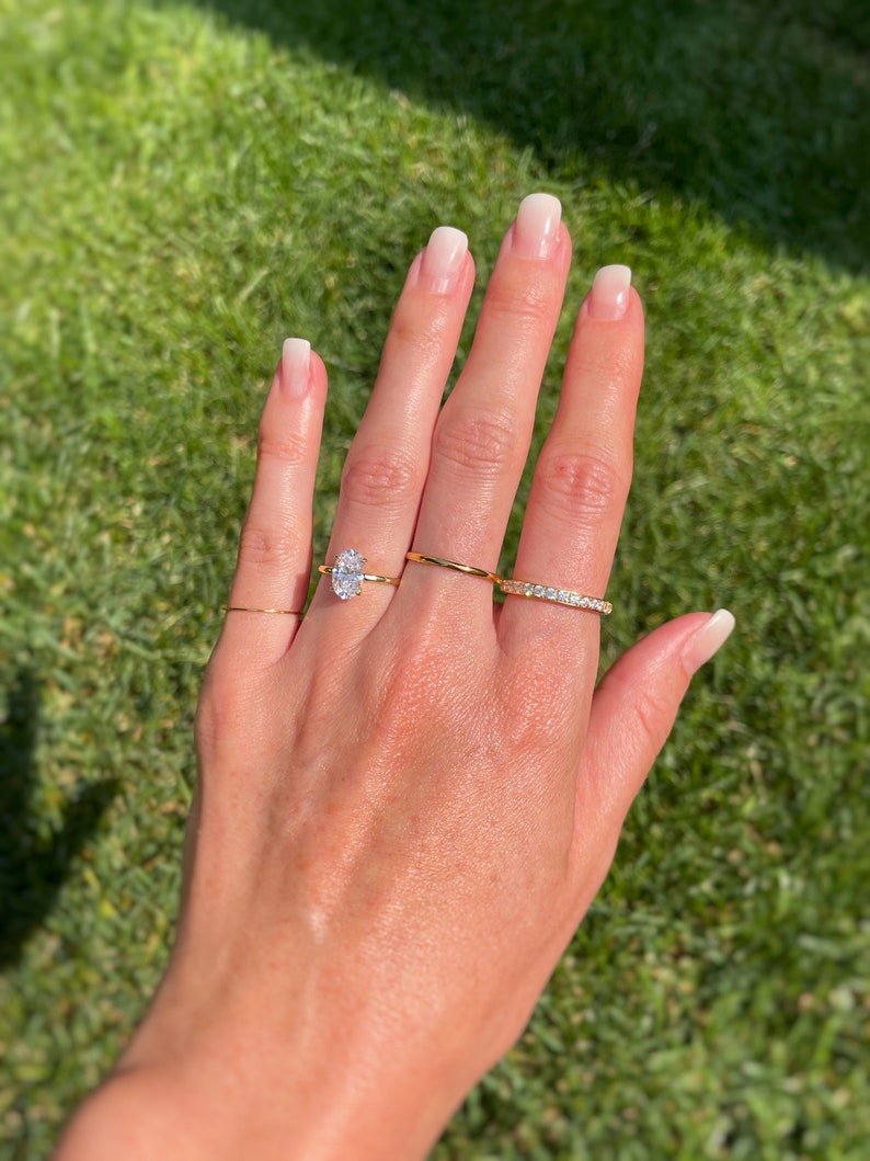 K&ouml;nnte beinhalten: Ein Set aus vier Goldringen an den Fingern. Ein Ring hat einen markanten oval geschliffenen klaren Stein, ein weiterer ist ein d&uuml;nner schlichter Goldring, der dritte ist ein einfacher Goldring und der vierte ist ein mit kleinen klaren Steinen besetzter Ring.